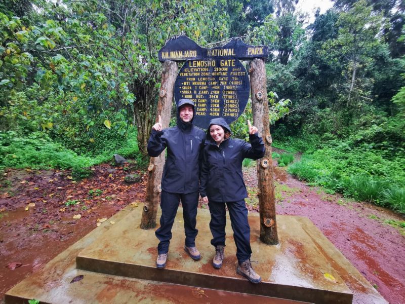 Two trekkers in front of the Lemosho sign and in wet gear as it's raining
