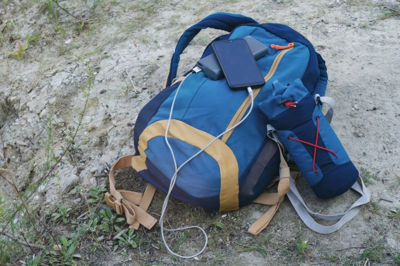 Smartphone being charged with portable powerbank, both resting on a backpack on the ground