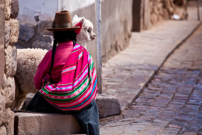 Woman in traditional clothes with alpaca sitting on stone in Cusco - Peru