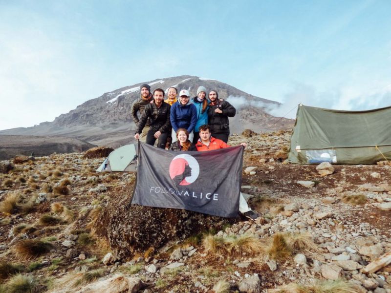 Group holding Follow Alice flag while standing in a campsite on Kilimanjaro