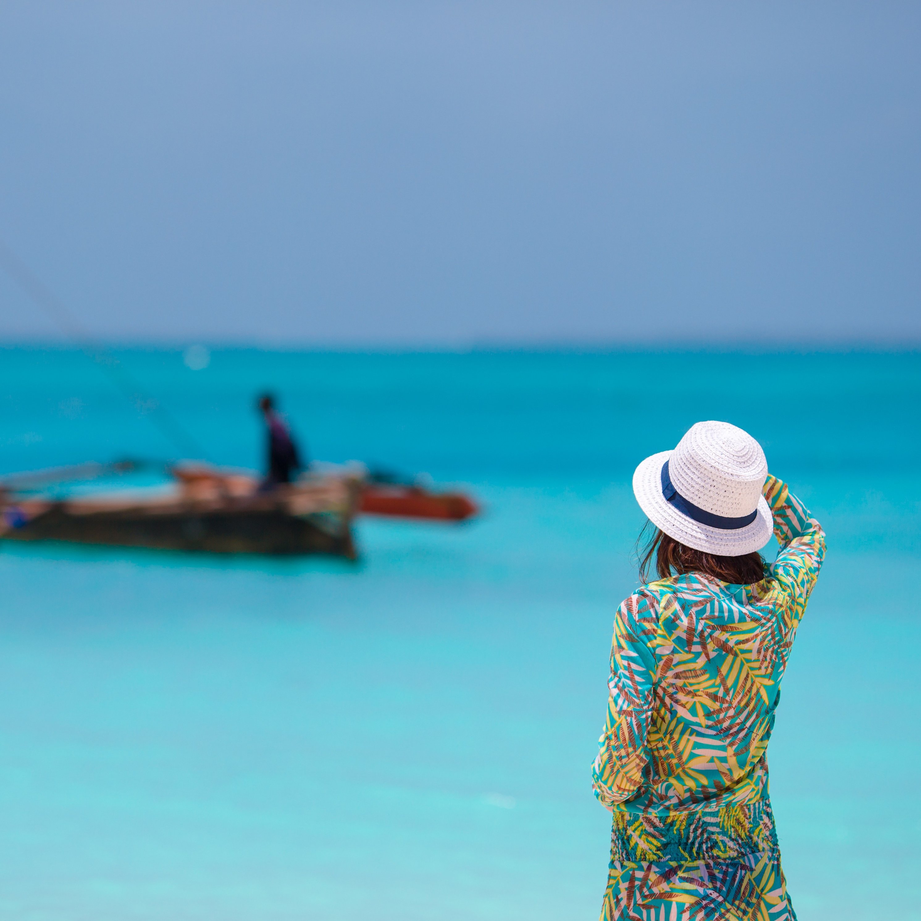 Female tourist in sunhat looking out over blue ocean towards dhow boat in Zanzibar