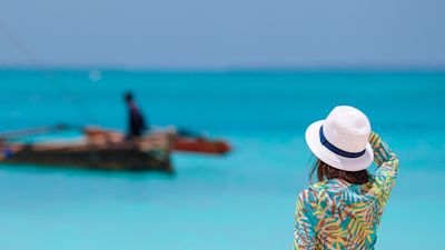 Female tourist in sunhat looking out over blue ocean towards dhow boat in Zanzibar