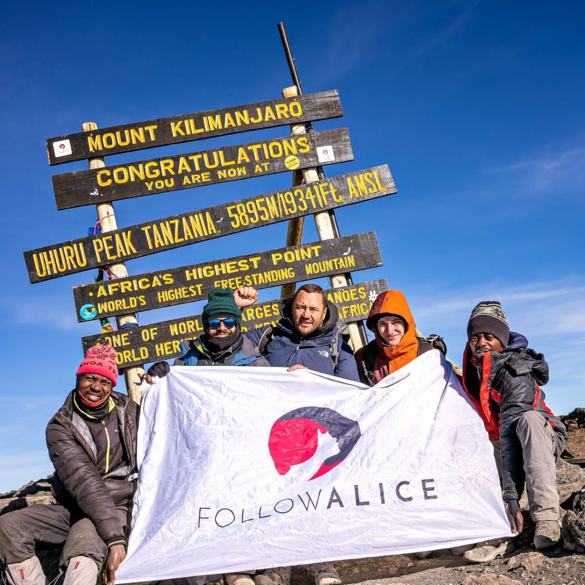 Group photo of trekkers sitting in front of Uhuru Peak sign and holding Follow Alice flag