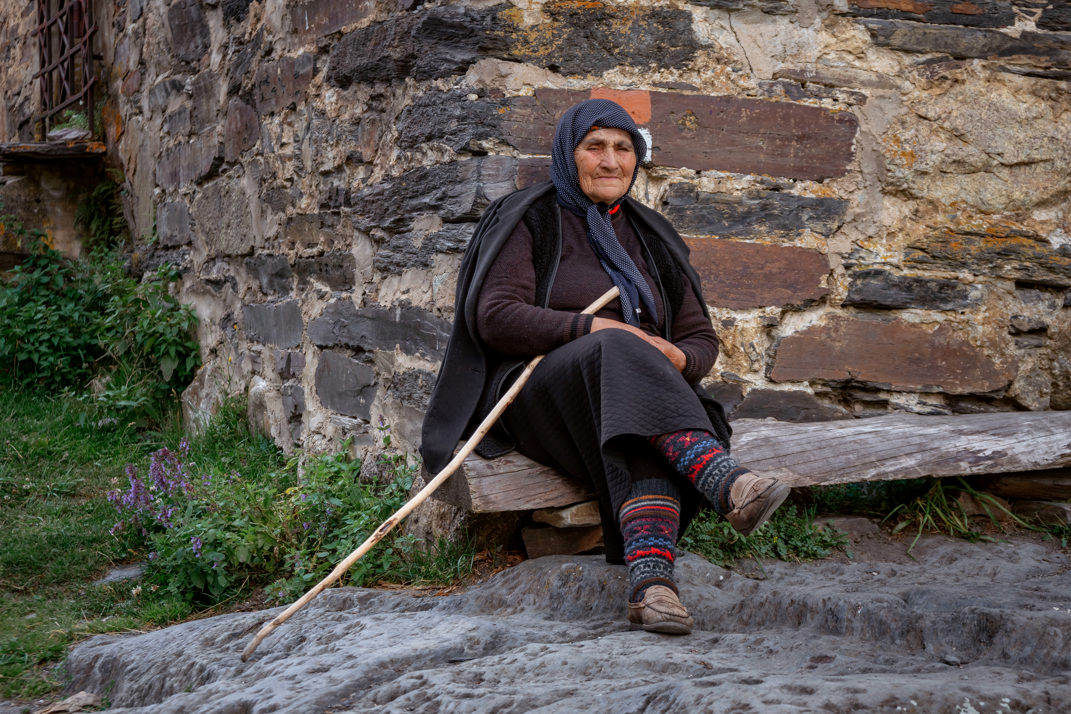 An elderly woman sits in front of the Svan tower with a wooden cane