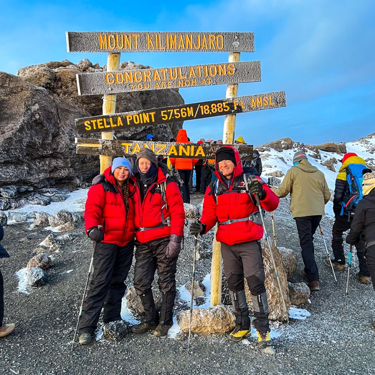Three climbers of different ages in Follow Alice down jackets at Stella Point sign on Mt Kilimanjaro trek