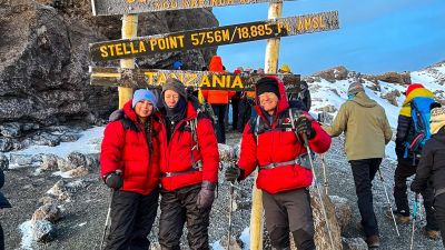 Three climbers of different ages in Follow Alice down jackets at Stella Point sign on Mt Kilimanjaro trek