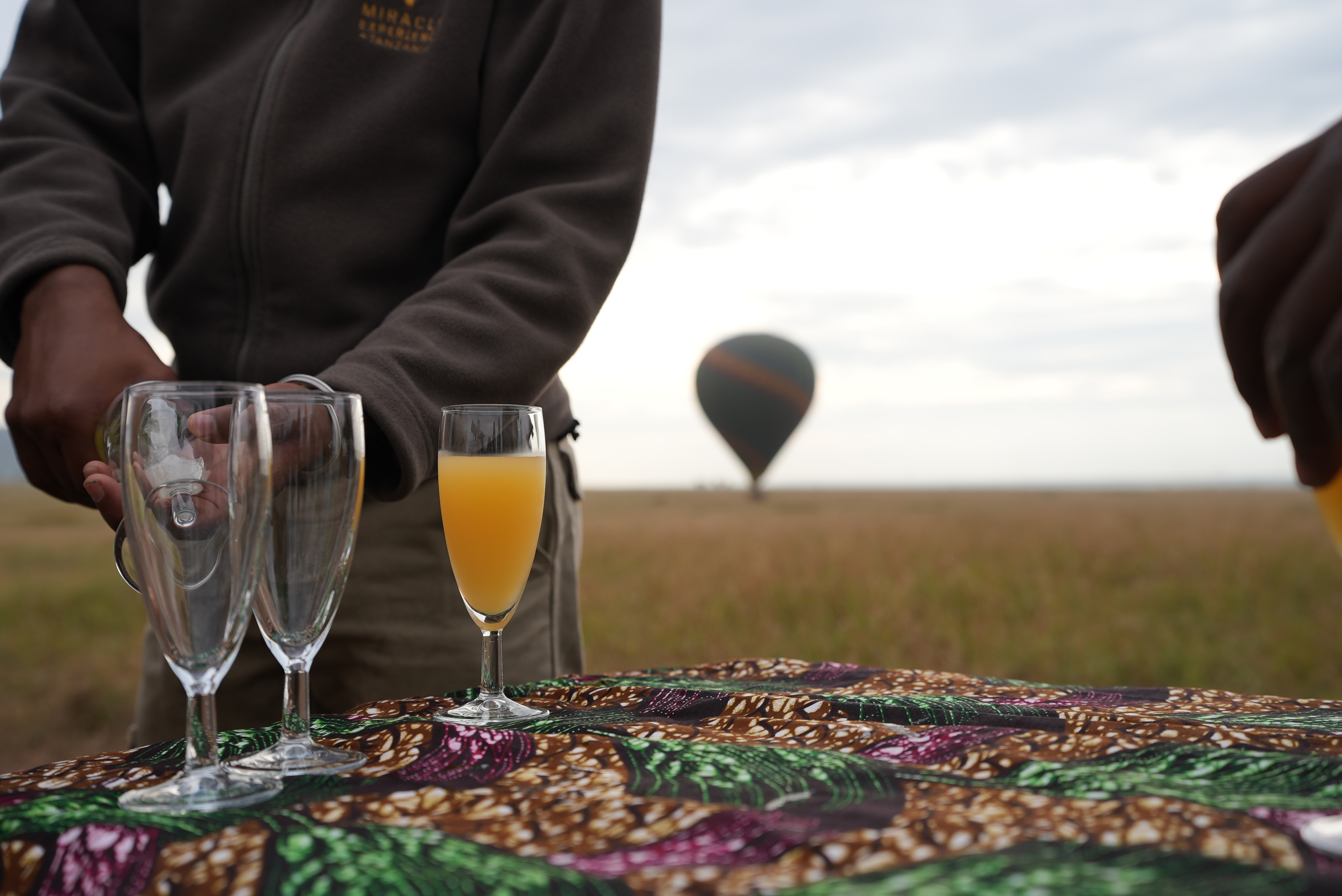 Man pouring mimosas in foreground with hot air balloon in background in Serengeti, Tanzania, April 2024