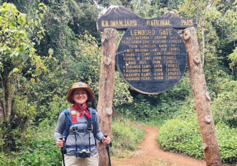 Smiling female hiker with film camera hanging around her neck standing at the Lemosho Gate sign on Kilimanjaro, August 2022