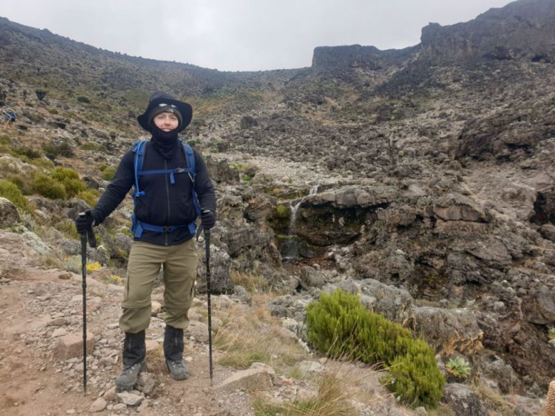 A male Kilimanjaro trekker stands in the moorland zone of Kilimanjaro with a small waterfall behind him