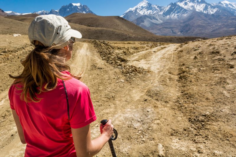 Female trekker in pink shirt and sports cap looking at barren trail on Annapurna Ciruit with mountain peaks in distnance