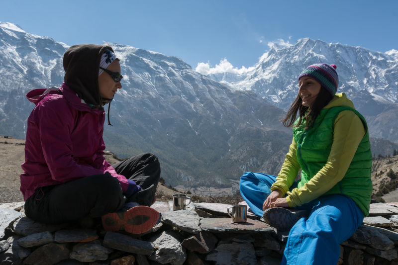 Two female trekkers seated and drinking hot drinks with Annapurna Mountains behind