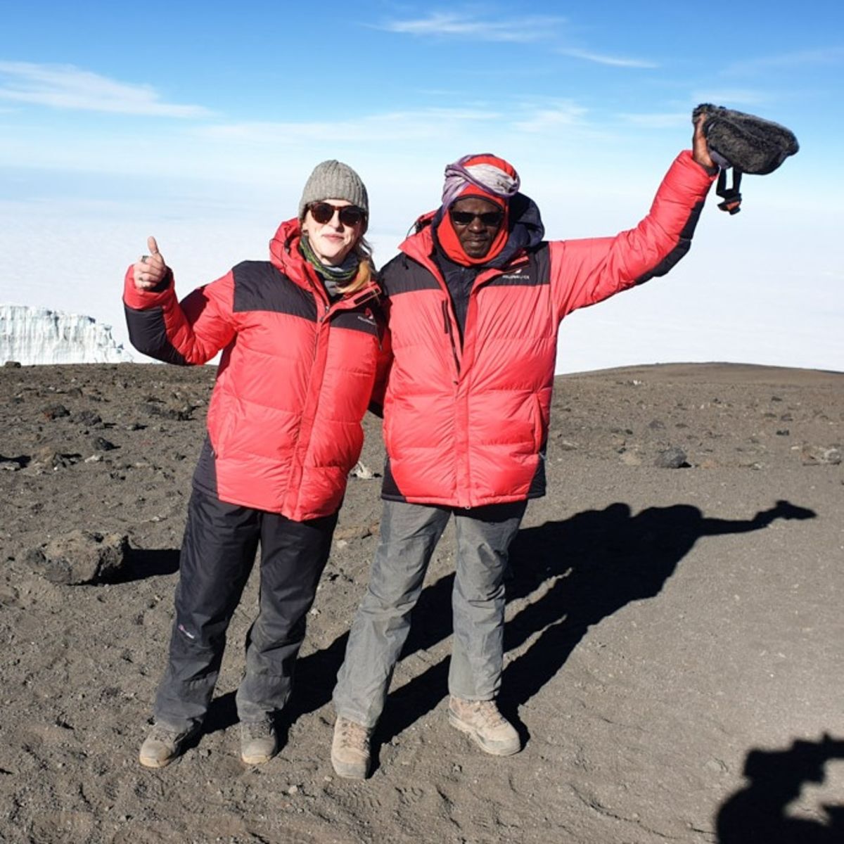 Tash and Chris at summit of Kilimanjaro on a clear, sunny day and waving at the camera with the glacier behind them