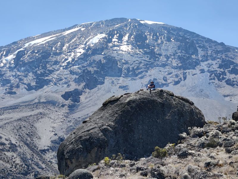 Man sitting on huge rock with Uhuru Peak behind him on Kilimanjaro climb