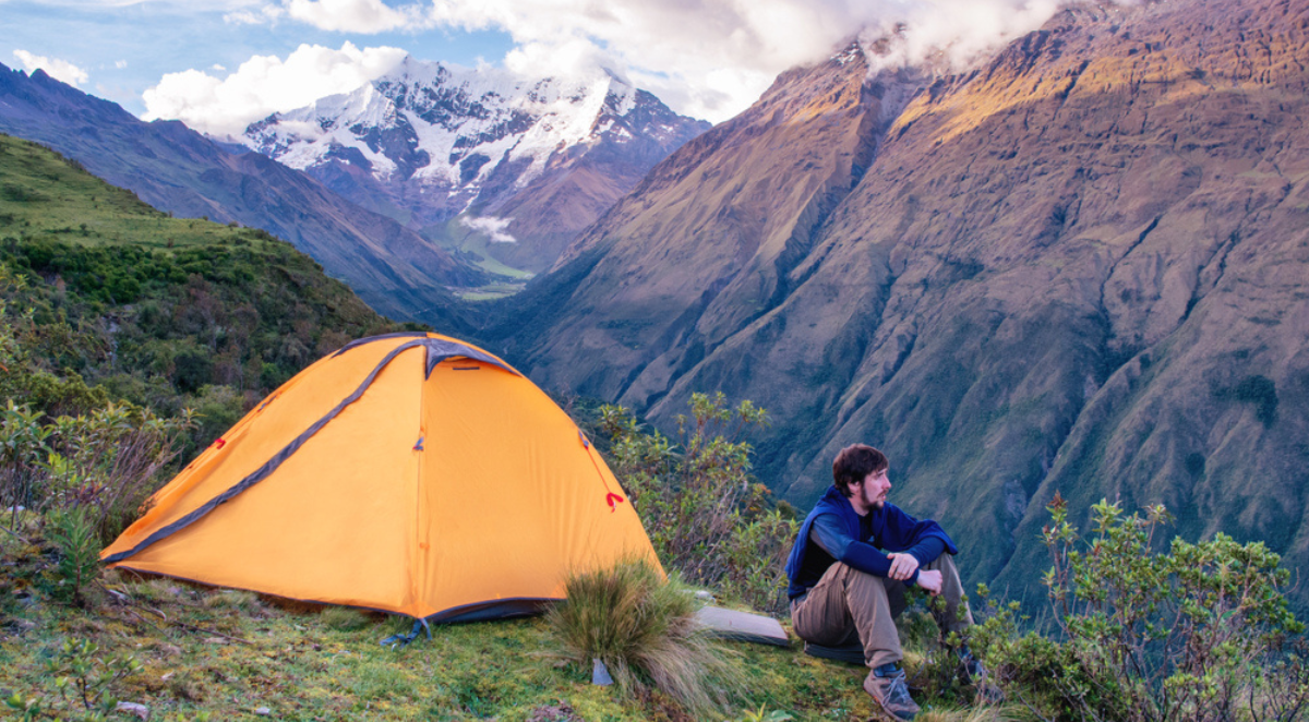 Male trekker seated next to small tent on Salkantay Trek looking at mountain view.