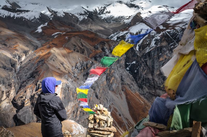 Female trekker wearing blue suncap standing near prayer flags and dramatic alpine mountains on Annapurna circuit in Nepal