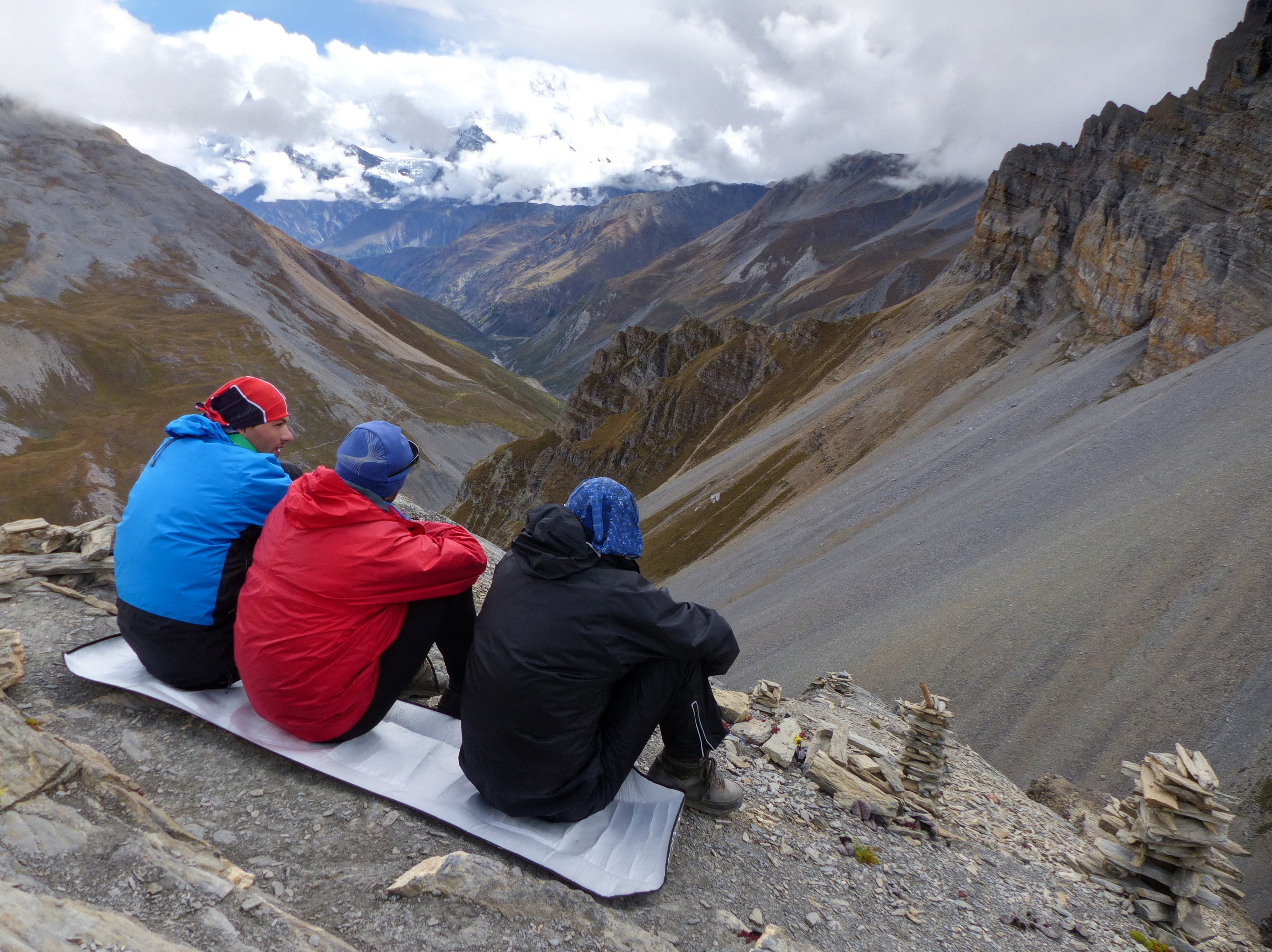 Three hikers sit on a ridge at Thorong High Camp, gazing down at the expansive Annapurna Mountain Range
