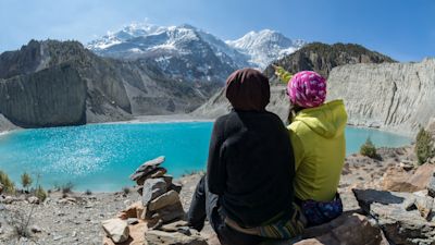 Two female hikers sit on the shore of Gangapurna lake and admire the summit of Gangapurna on the Annapurna Circuit, in Manang