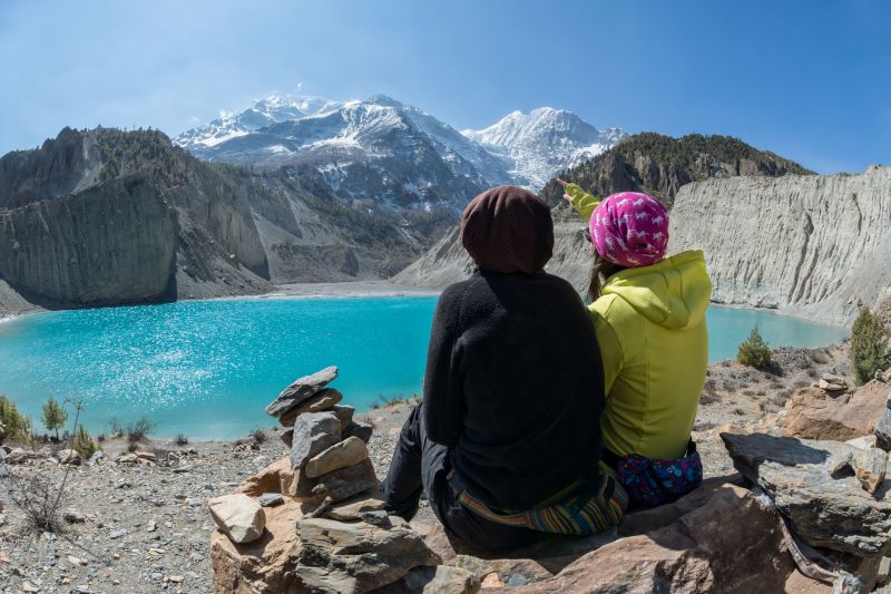Two female hikers sit on the shore of Gangapurna lake and admire the summit of Gangapurna on the Annapurna Circuit, in Manang