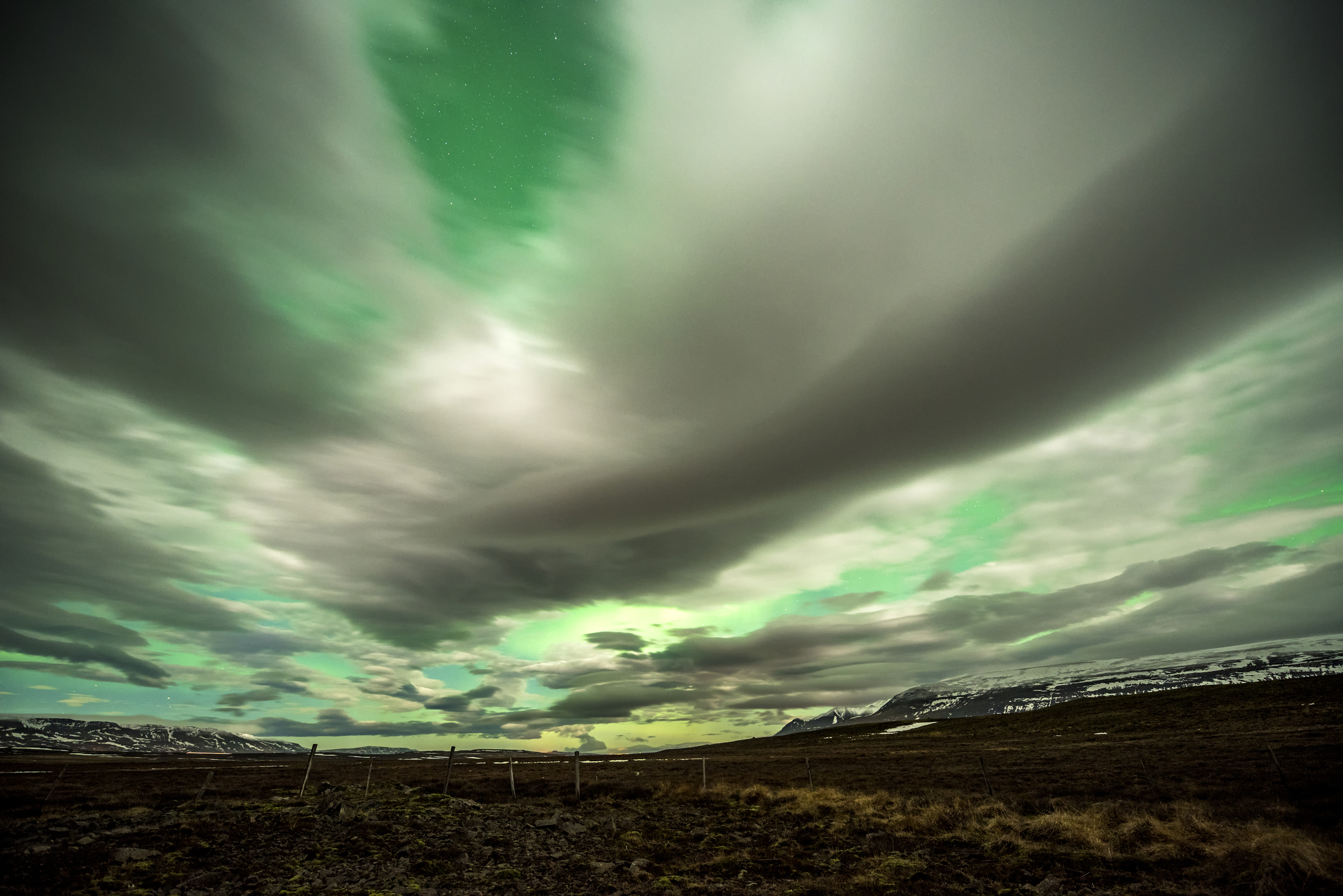 Aurora Borealis glows over a frozen lake surrounded by low cloud cover and stars