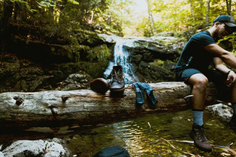 A hiker sits on a log that's balanced high across a stream and has a pair of hiking boots and socks placed on the log beside him