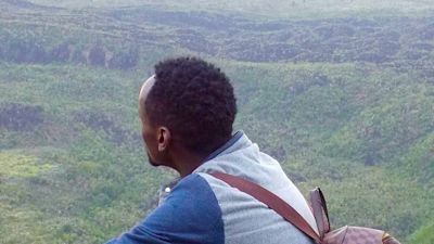 Young African man in yellow boots and wearing a checked backpack sitting on rock high above green landscape