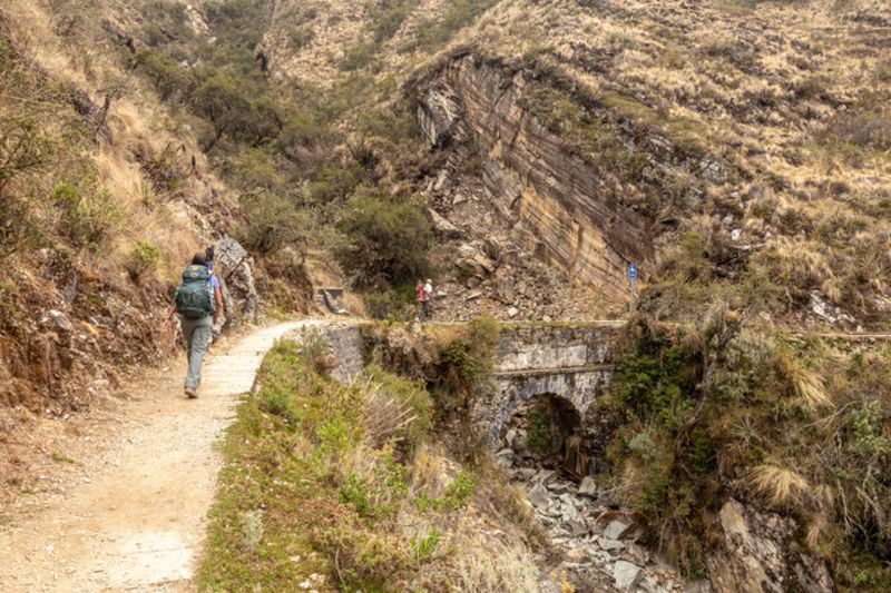 Trekkers on a flat dirt contour path on the Salkantay Trek approaching a small stone bridge 