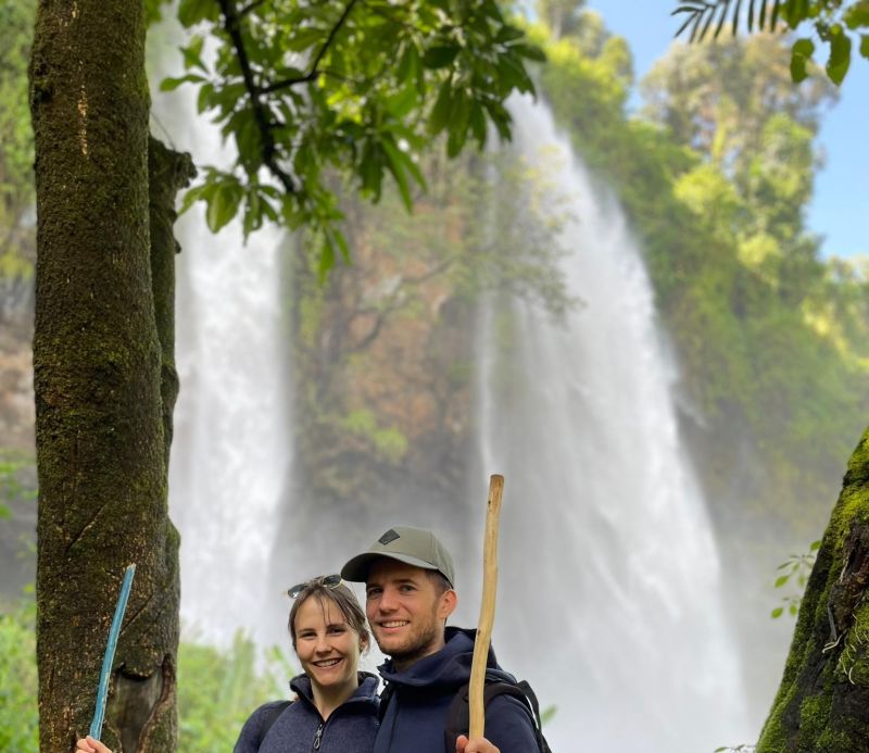 Male and female hiker with hiking staffs standing in front of Sipi Falls in Uganda, August 2022