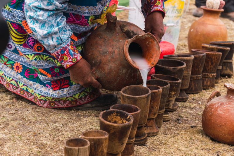 Woman in traditional dress using ceramic pot of traditional chicha de jora and wooden glasses in a traditional ritual 