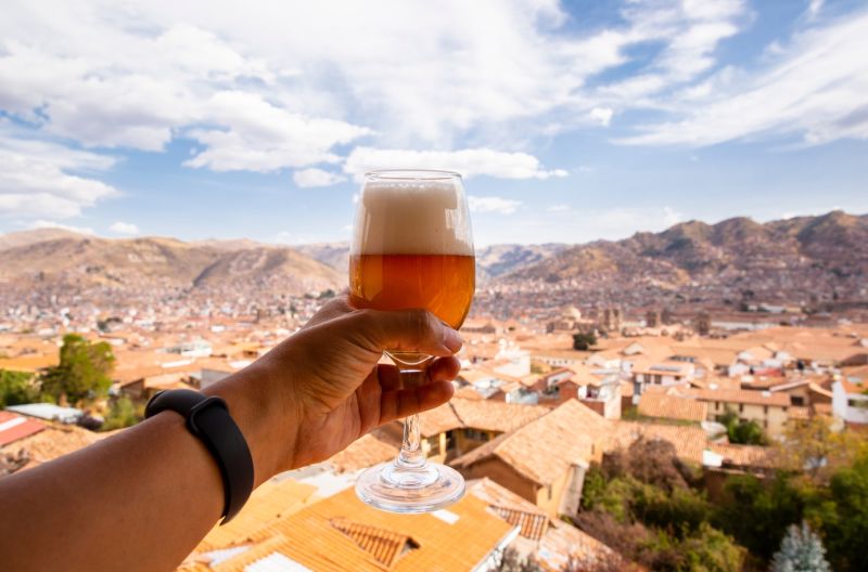 Man-s hand holding beer (cerveza) in glass in salute to cityscape of Cusco below, Peru 