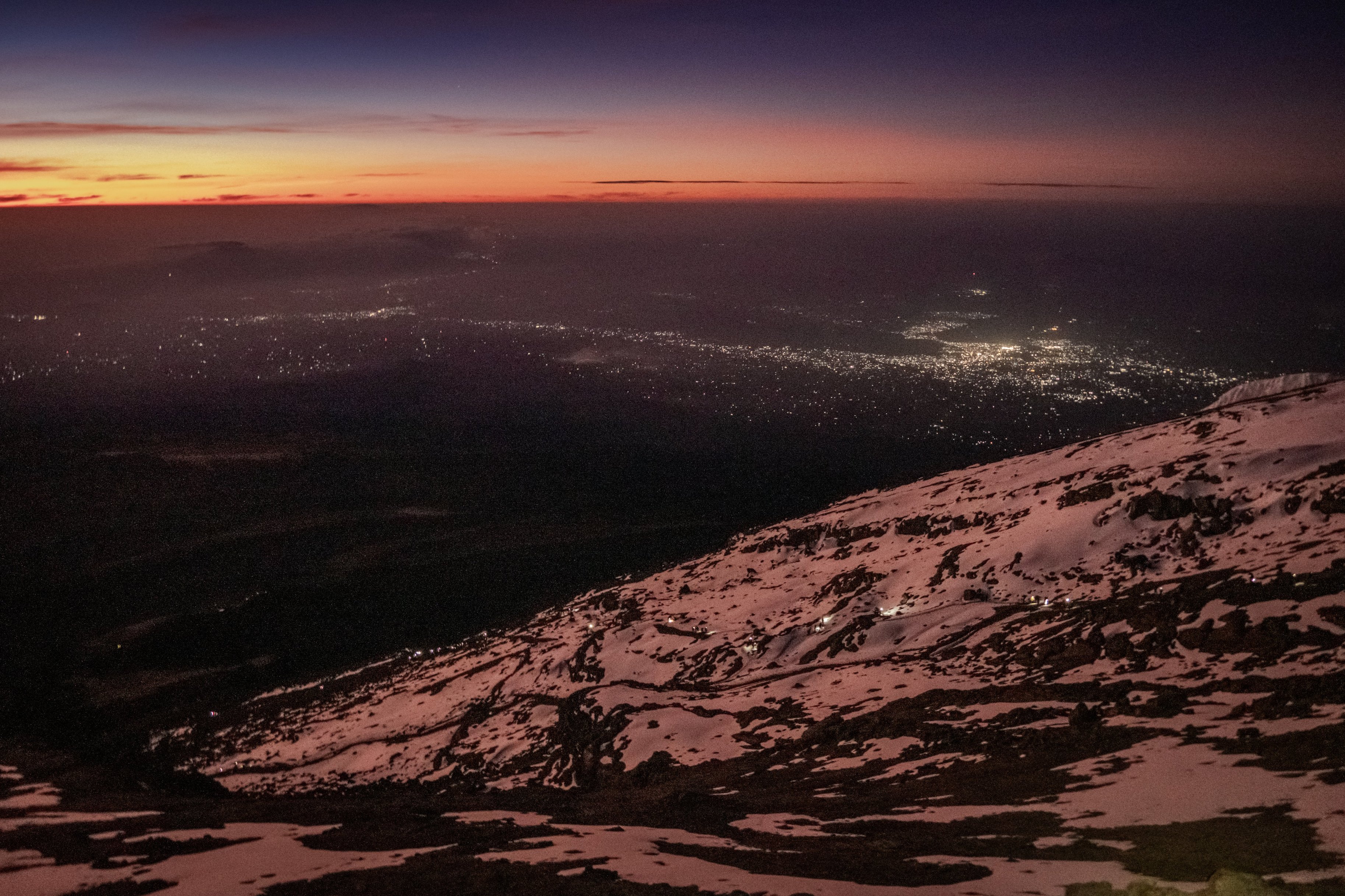 Thin line of trekkers with headlamps showing against snowy slope of Kilimanjaro, climbing to Uhuru Peak on summit night, lights of Moshi below and dawn sky