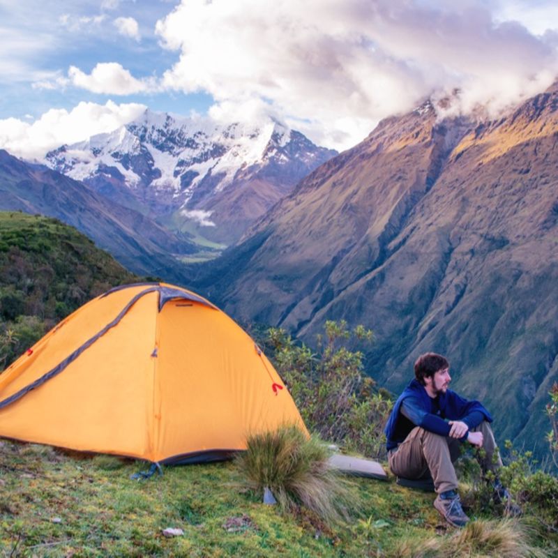 Male trekker seated next to small tent on Salkantay Trek looking at mountain view. Peru