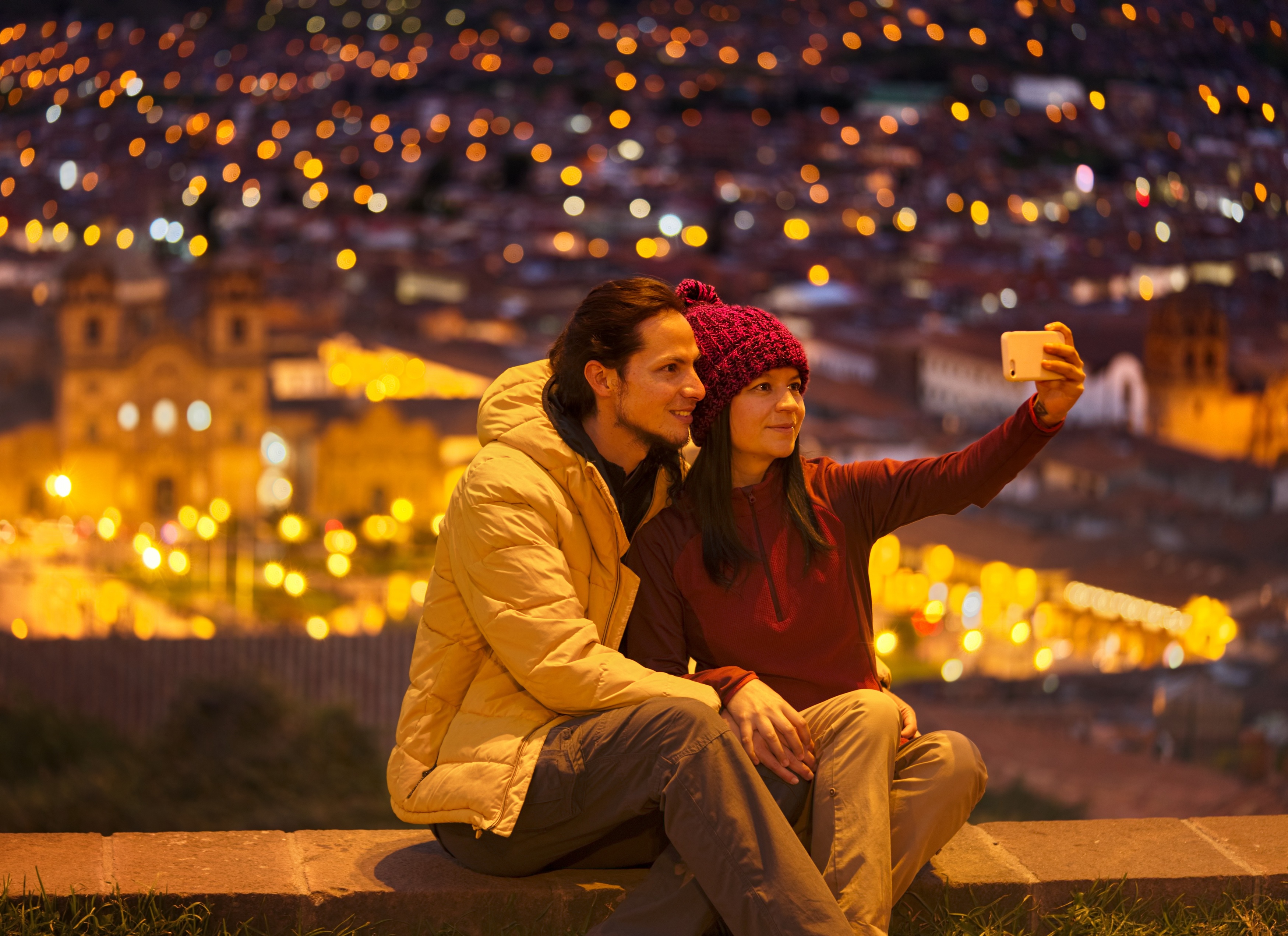 Couple taking a selfie at night while seated on a low wall with the lights of central Cusco city below them, Peru
