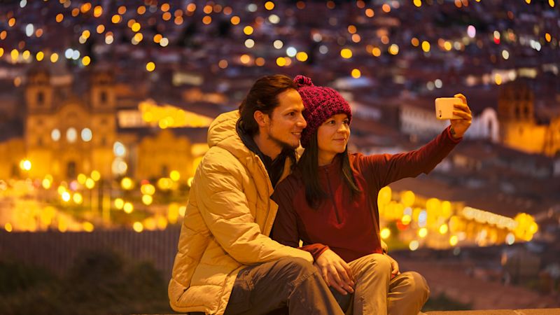 Couple taking a selfie at night while seated on a low wall with the lights of central Cusco city below them, Peru