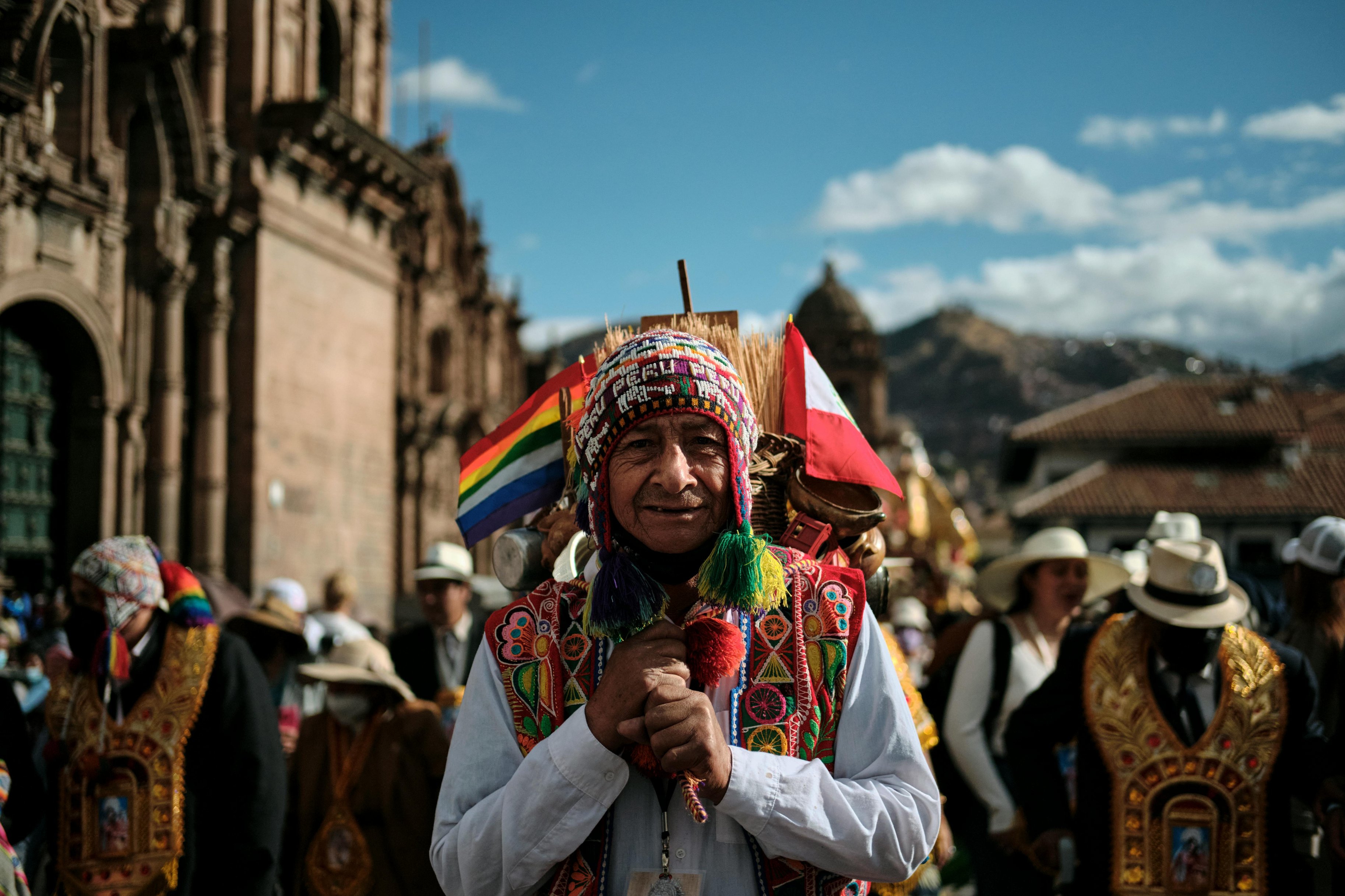 Man in traditional woven hat and traditional dress holding celebratory flags while standing in a busy plaza by the cathedral in Cusco on a festival day, Peru