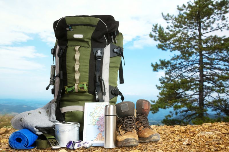 Large green backpack with hiking boots, flask, sleeping roll and map on ground outside