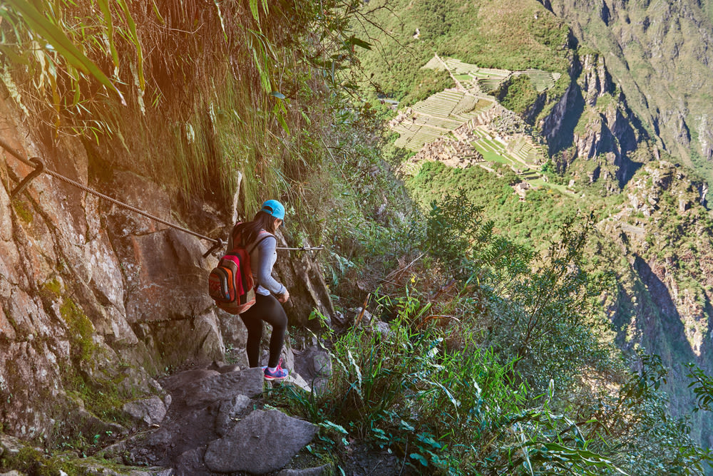 Young woman trekker in blue cap walking down steep stone steps and holding onto a rail on a trail to Machu Picchu, with ruins in distance