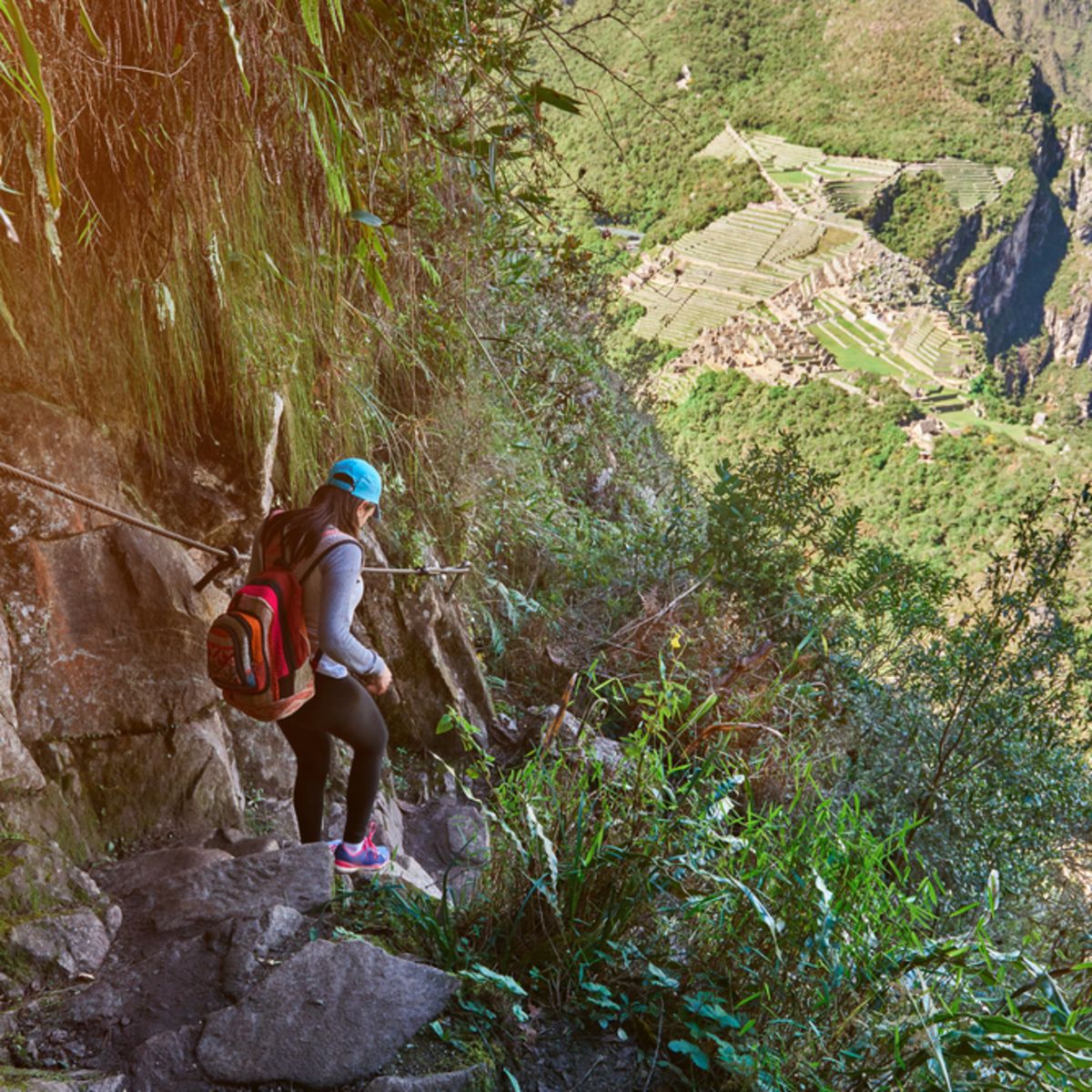 Young woman trekker in blue cap walking down steep stone steps and holding onto a rail on a trail to Machu Picchu, with ruins in distance