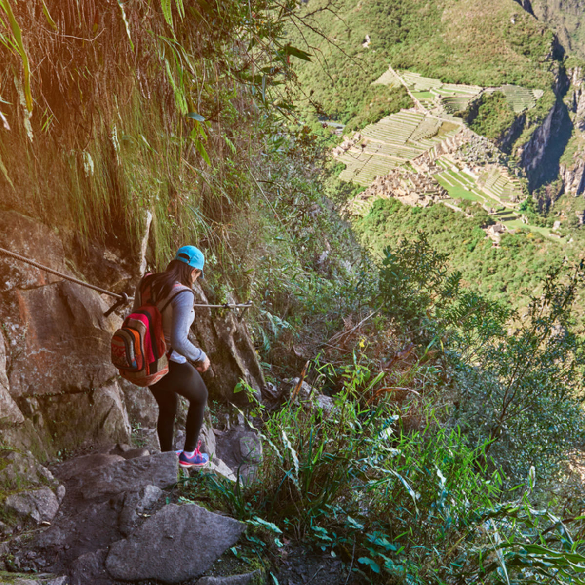 Young woman trekker in blue cap walking down steep stone steps and holding onto a rail on a trail to Machu Picchu, with ruins in distance