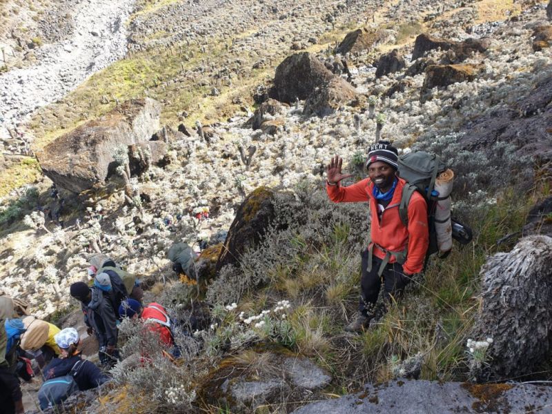 Man in red jersey waving at the camera as a group of hikers climbs down Barranco Wall on Kilimanjaro