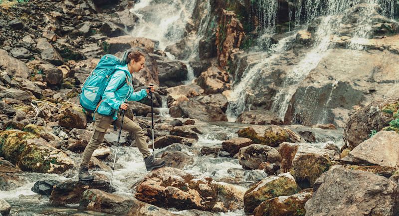 Woman hiker walking across rocky stream using trekking poles and carrying rucksack