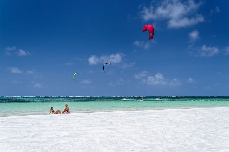 Two women in bikinis sitting in the shallows of a Kenya beach with three windsurfers on the water