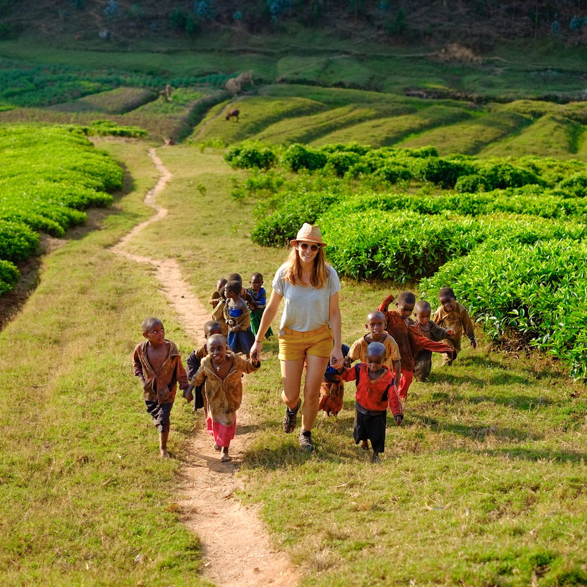 A tourist holding hands with children walking up a hill in a tea plantation on a dusty path in Rwanda