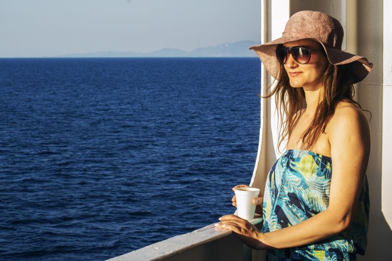 Woman in sunhat and sunglasses holding a coffee cup and looking out from a ship at the ocean