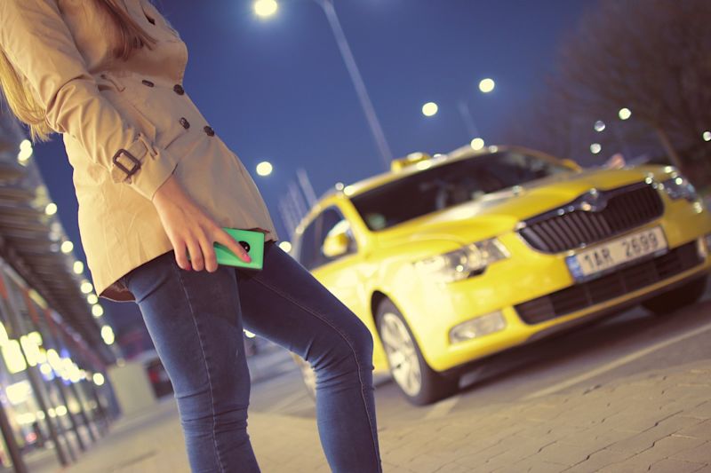 Woman holding phone on city street being approached by yellow taxi