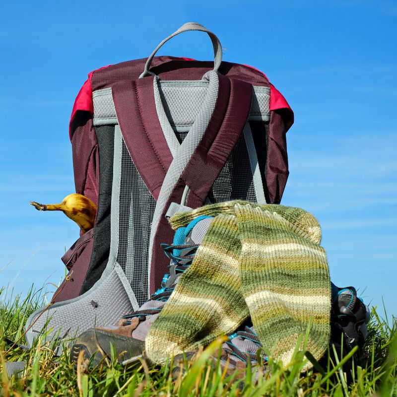 Maroon-and-grey backpack with banana poking out of side pocket and hiking boots with striped green socks placed on the grass