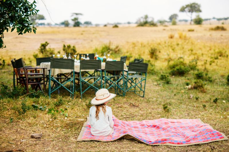 Little girl in hat seated on Maasai blanket by bush lunch table, Kenay==ya game reserve safari