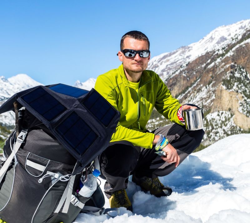 Male trekker crouched down in the snow, backpack open, sunglasses on, and holding a hot drink