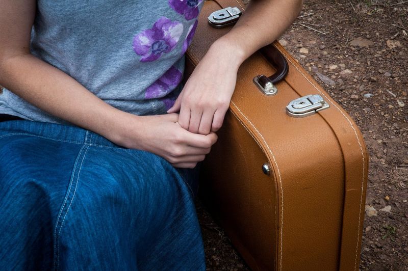 Close up of girl in denim skirt leaning on tan suitcase, seated on ground
