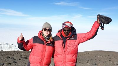 Tash and Chris at summit of Kilimanjaro on a clear, sunny day and waving at the camera with the glacier behind them