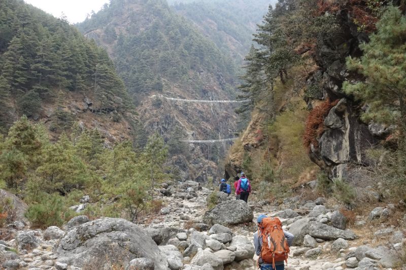 Hikers wearing backpacks walking over rocks towards a suspension bridge in a forest valley along the Everest Base Camp trek route